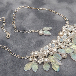 May include: A silver chain necklace with a cluster of iridescent white and green glass beads and leaves. The necklace is on a grey background.