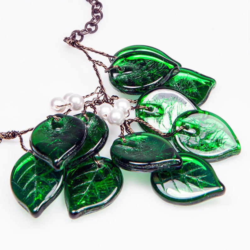 May include: A green glass leaf necklace with a brown chain and white pearl accents. The leaves are made of textured glass and have a metallic sheen.