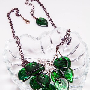 May include: A green glass leaf necklace with a silver chain and white pearl accents. The necklace is displayed on a clear glass dish.
