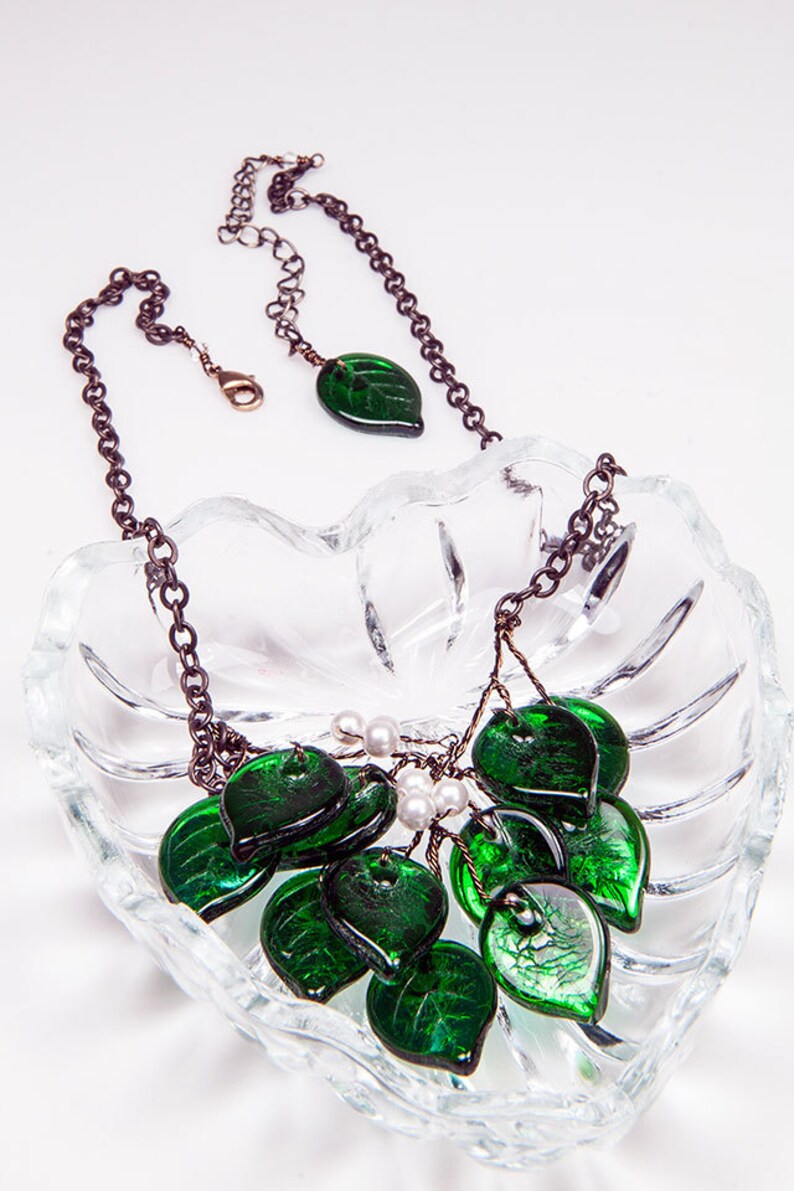 May include: A green glass leaf necklace with a silver chain and white pearl accents. The necklace is displayed on a clear glass dish.
