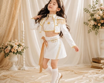 May include: A white two-piece outfit consisting of a cropped jacket with gold fringe epaulettes and a matching skirt with a gold ribbon sash. The jacket features gold trim and a high collar. The skirt includes a gold waist detail and a side ribbon.
