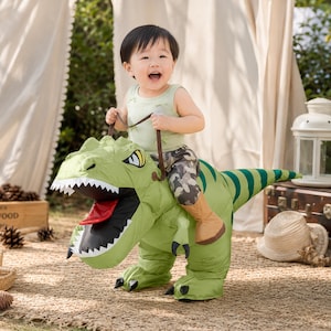 May include: A green inflatable dinosaur costume featuring a large head with sharp white teeth, a red tongue, and dark green stripes along the tail. The costume includes brown straps and tan boot covers, designed to be worn around the waist.