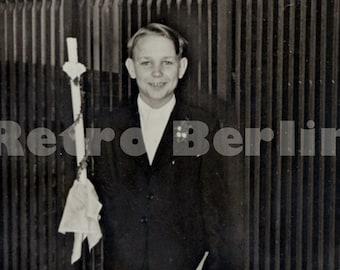 May include: A young boy in a suit and tie holds a white candle in his right hand. He is standing in front of a black metal gate. The photo is in black and white.