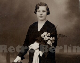 May include: A black and white photo of a woman in a dark dress holding a bouquet of white flowers. She is standing next to a small table with a white tablecloth.