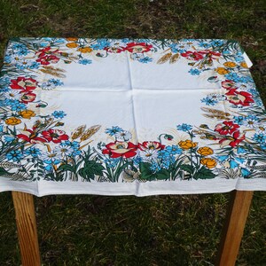 Pode incluir: Toalha de mesa branca com uma borda floral com papoilas vermelhas, centáureas azuis, botões-de-ouro amarelos e folhagem verde. A toalha de mesa está sobre uma mesa de madeira ao ar livre, com um fundo gramado.
