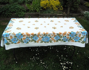 May include: White tablecloth with a blue and orange floral pattern. The tablecloth is laid out on a table with black metal legs. The table is set on a grassy lawn.