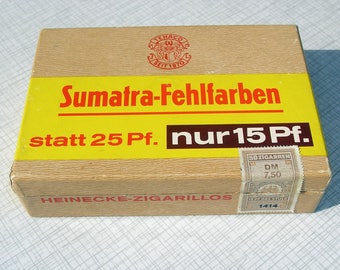 May include: A brown cardboard box with a yellow label that reads "Sumatra-Fehlfarben statt 25 Pf. nur 15 Pf." The box also has a brown stamp with the text "50 ZIGARREM DM 7,50" and "TRAF DAS STUER 1414". The box is labeled "HEINECKE-ZIGARILLOS".