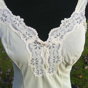May include: Cream-colored slip with a V-neckline and delicate floral lace trim. The lace is a soft, off-white color and is arranged around the neckline and straps. A small bow accents the center of the neckline.