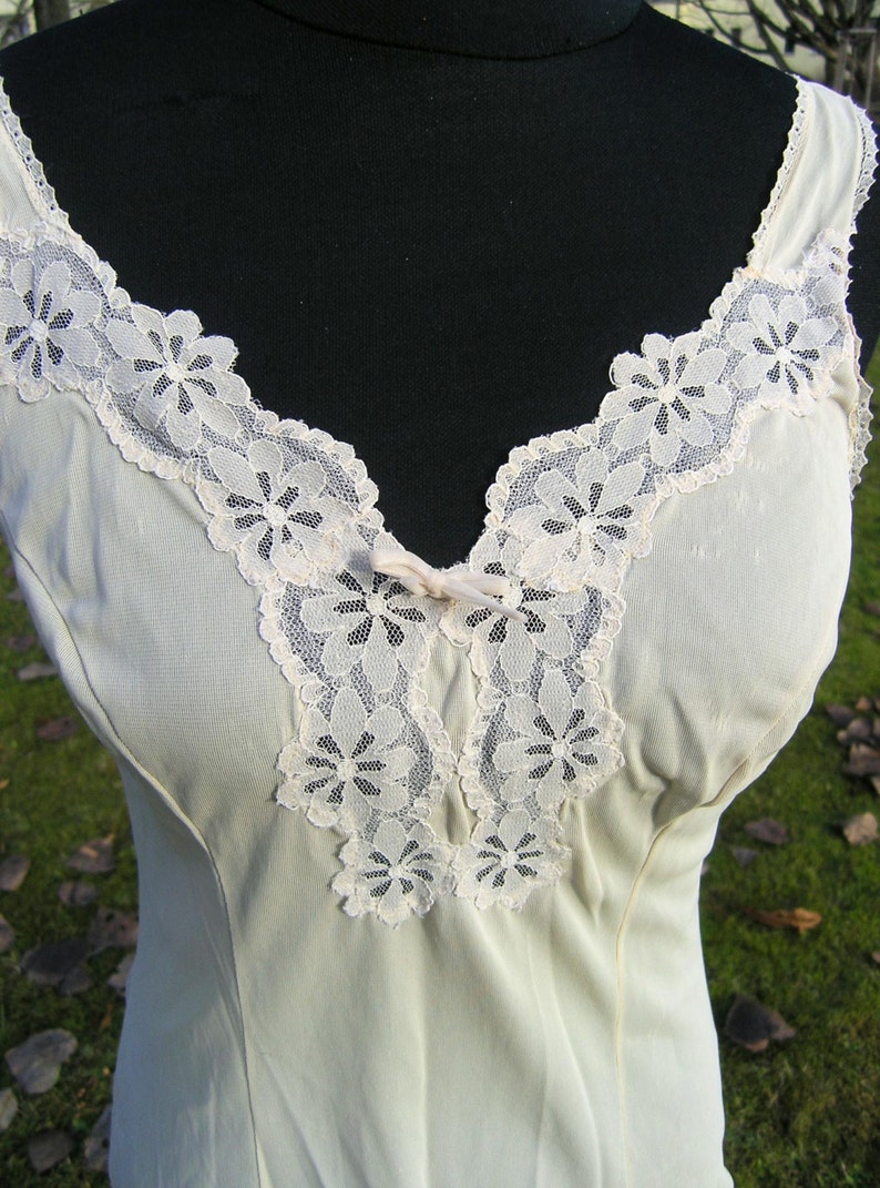 May include: Cream-colored slip with a V-neckline and delicate floral lace trim. The lace is a soft, off-white color and is arranged around the neckline and straps. A small bow accents the center of the neckline.