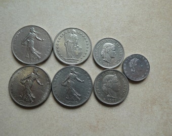 May include: A collection of seven silver coins featuring different designs and dates. The coins are arranged in a semi-circle on a light brown surface.