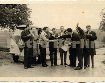 May include: A group of men standing around a vintage white Volkswagen van with the license plate "S-HR 24". Some of the men are holding drinks and talking. The van is parked in front of a grassy field.