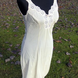 May include: Cream-colored vintage slip dress on a black mannequin. The dress features a V-neckline and straps, both trimmed with delicate white lace. The hemline also has a lace trim. The dress is displayed outdoors.