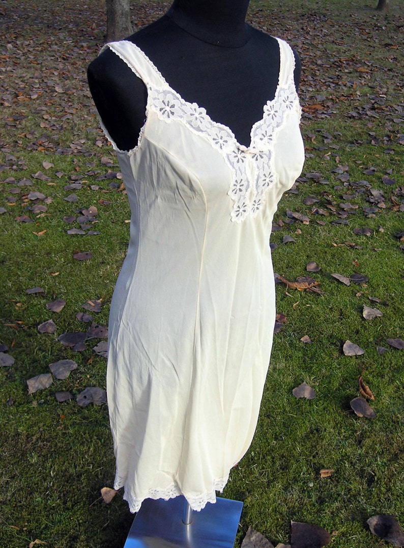 May include: Cream-colored vintage slip dress on a black mannequin. The dress features a V-neckline and straps, both trimmed with delicate white lace. The hemline also has a lace trim. The dress is displayed outdoors.