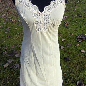 May include: A cream-colored slip dress with a V-neckline and delicate lace trim. The dress features a fitted silhouette and thin straps. The lace detailing is present on the neckline and hem, adding a touch of elegance to the garment.