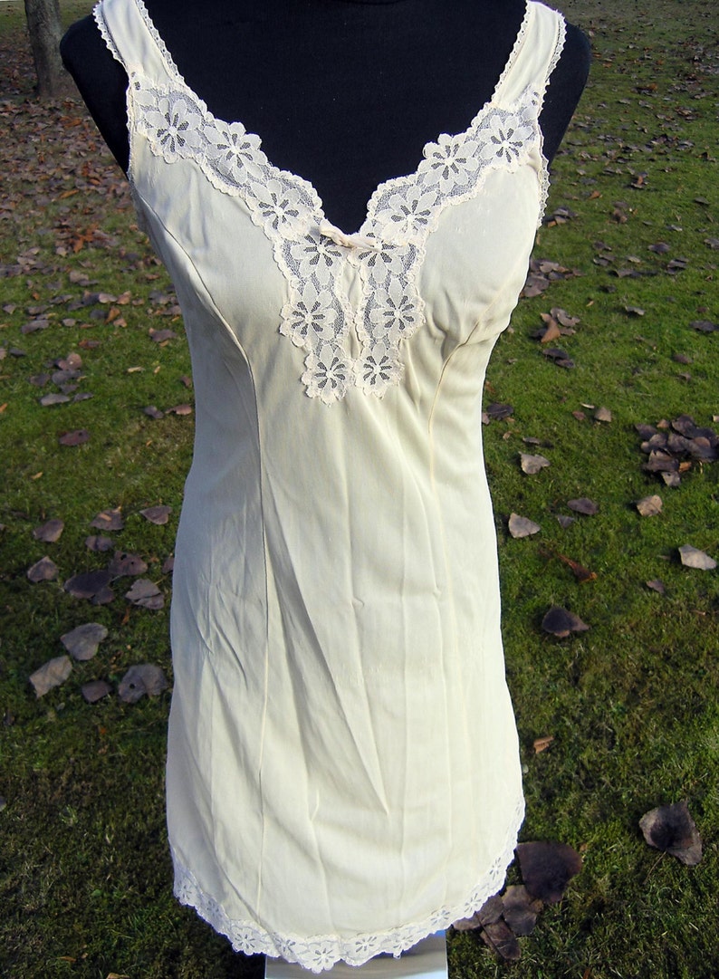 May include: A cream-colored slip dress with a V-neckline and delicate lace trim. The dress features a fitted silhouette and thin straps. The lace detailing is present on the neckline and hem, adding a touch of elegance to the garment.