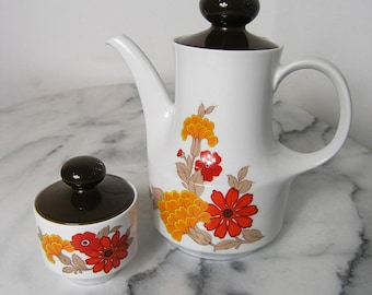 May include: A white ceramic teapot and sugar bowl with brown lids and floral designs. The teapot has a brown handle and a spout. The sugar bowl has a lid with a brown knob. The flowers are orange, red, and white.
