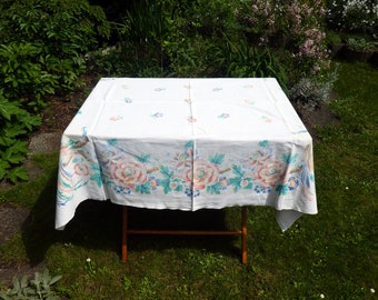 May include: A white tablecloth with a floral pattern in shades of pink, blue, and green. The tablecloth is laid out on a wooden table and is surrounded by green grass.