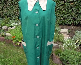May include: A sleeveless, emerald green apron-style garment with a cream-colored, plaid-patterned collar and pocket accents. The apron has a button-down front and two front pockets. The garment is displayed on a mannequin in a garden setting.
