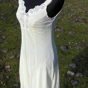 May include: A cream-colored slip dress with delicate lace trim along the neckline, straps, and hem. The dress is sleeveless and has a V-neckline. The fabric appears lightweight and flowing, suggesting a vintage or romantic style.