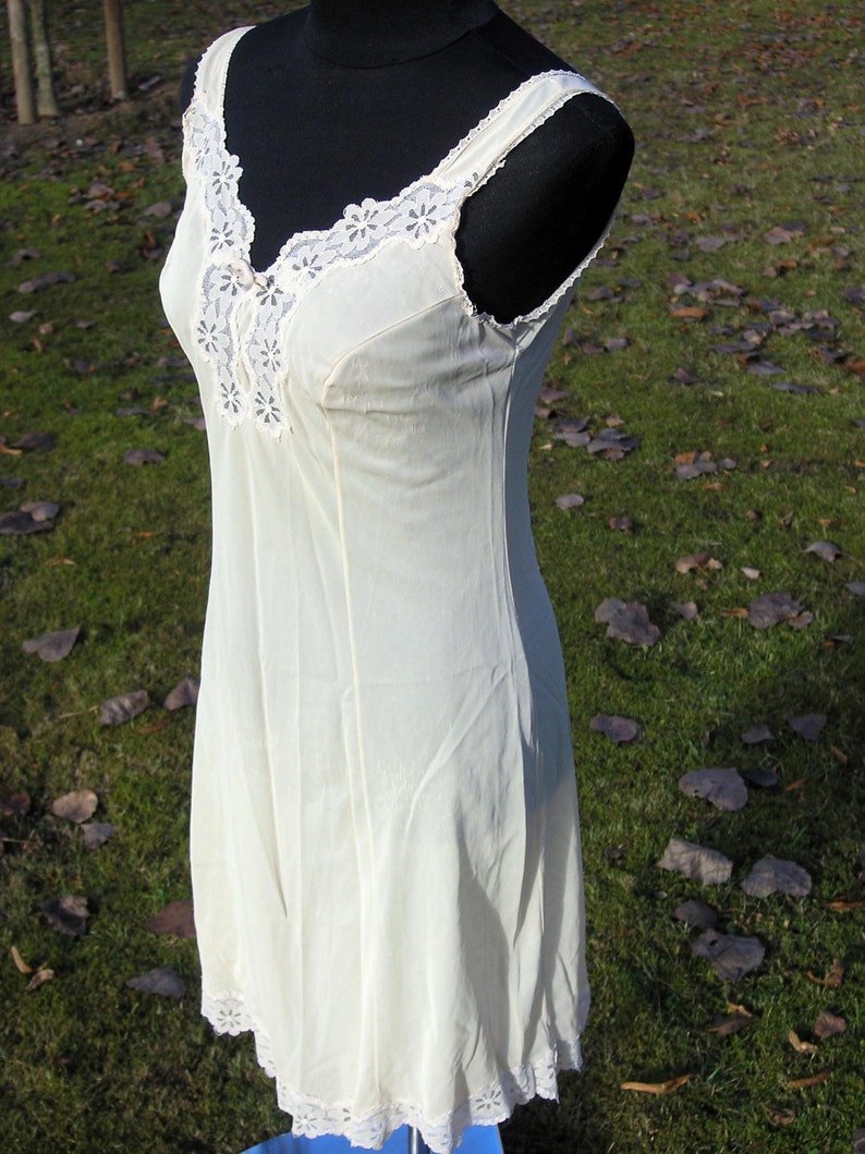 May include: A cream-colored slip dress with delicate lace trim along the neckline, straps, and hem. The dress is sleeveless and has a V-neckline. The fabric appears lightweight and flowing, suggesting a vintage or romantic style.