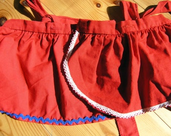 May include: A red apron with white lace trim and blue scalloped edging. The apron has two straps and a gathered waist.