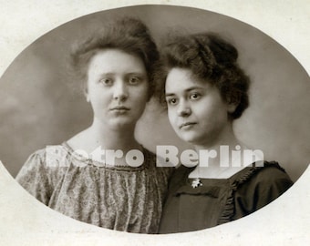 May include: A black and white vintage photograph of two young women with their hair styled in updos. The women are wearing dresses and are looking at the camera. The photo is oval shaped and has a white border.
