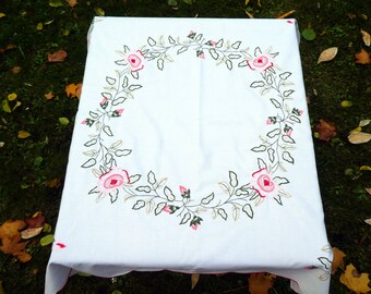 May include: White tablecloth with an embroidered floral design. The design features a wreath of pink roses and green leaves.