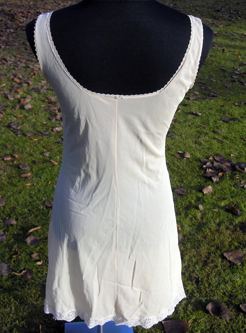 May include: Cream-colored slip dress with a scoop neckline and delicate lace trim. The sleeveless dress is made of a soft, flowing fabric and features a subtle seam down the back. The hemline is also trimmed with lace.