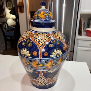 May include: A large, round ceramic jar with a matching lid, featuring a glossy finish. The surface is decorated with intricate hand-painted patterns in deep blue, vibrant orange, yellow, and white, including floral motifs and geometric grid designs.