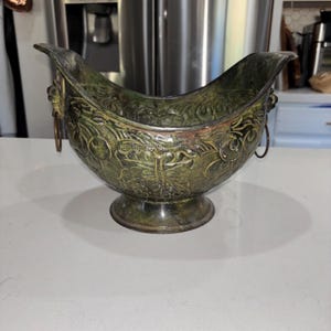 Puede incluir: Un cuenco de metal en tonos verde oscuro y bronce con una base de pedestal y una parte superior curva y abierta. El exterior presenta un intrincado patrón floral en relieve y dos acentos de cabeza de león en los lados, cada uno con un anillo.