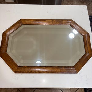 May include: An octagonal wall mirror featuring a medium-toned brown wood frame with a visible grain pattern. The mirror glass has a beveled edge that follows the eight-sided shape of the frame, creating a reflective surface with depth and dimension.