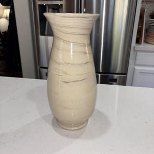 Puede incluir: Un jarrón de cerámica alto y acampanado con una base lisa de color crema y patrones de mármol gris horizontales y ondulados. El recipiente tiene una boca ancha y abierta que se estrecha hacia un cuerpo redondeado y bulboso.