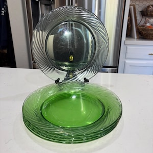 Puede incluir: Un juego de cuatro platos redondos de vidrio con un tono verde vibrante y un patrón de espiral giratorio que irradia desde el centro hacia el borde. Los platos están apilados, con uno de ellos colocado verticalmente sobre un soporte negro.