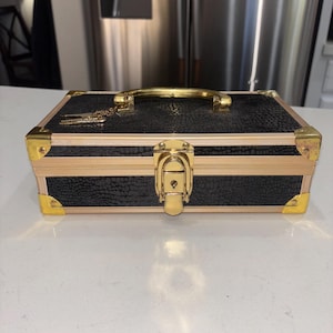 May include: A rectangular black storage case with a textured surface, featuring gold-toned metal corner reinforcements, a top handle, and a central locking latch. A small gold-toned charm hangs from the handle base on the top of the case.