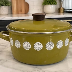 Peut inclure: Une marmite en métal émaillé vert olive avec un couvercle assorti et un bouton de préhension en bois brun foncé. La marmite possède deux poignées latérales et un motif géométrique circulaire blanc répété autour du corps extérieur.