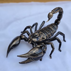 Pode incluir: Uma figura de escorpião metálico com acabamento em bronze escuro e preto, apresentando padrões gravados intrincados no corpo segmentado, cauda curva e pinças grandes. A escultura é retratada em uma postura defensiva com o ferrão erguido.