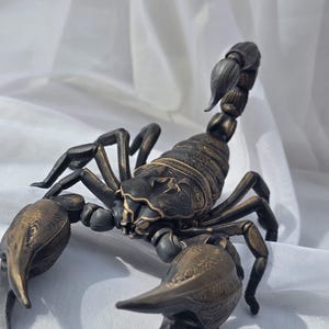 May include: A dark bronze-coloured scorpion figurine featuring intricate etched detailing on its segmented body, large pincers, and a curved tail. The sculpture displays a metallic finish with lighter highlights on the raised surfaces of its exoskeleton.
