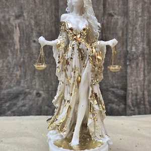 Könnte beinhalten: Eine weiße Frauenfigur mit langem Haar in einem fließenden Kleid, das mit Blattgoldakzenten verziert ist. Sie hält in jeder Hand eine kleine Waage. Die Figur steht auf einem runden weißen Sockel mit einem goldenen Waage-Sternzeichen vorne.