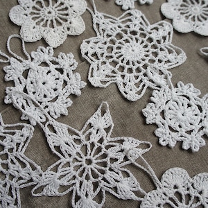 Puede incluir: Una colección de adornos blancos de copos de nieve, hechos a ganchillo, en varias formas y tamaños. Las decoraciones hechas a mano están dispuestas sobre una superficie de tono neutro, mostrando los intrincados detalles del trabajo de la lana. Ideal para la decoración navideña.