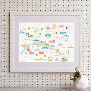 May include: A colorful illustrated map of Paris, France, with landmarks and neighborhoods labeled. The map is framed in a white frame and is hanging on a white pegboard wall.
