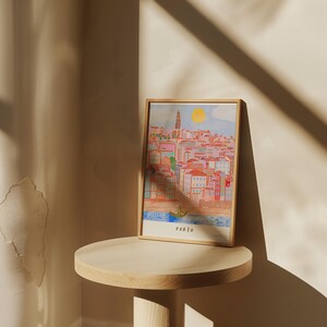 May include: A framed illustration of the city of Porto, Portugal. The illustration is in a colorful, cartoon style and depicts a cityscape with buildings, a river, and a boat. The frame is wooden and has a light brown finish. The text "PORTO" is printed at the bottom of the illustration.