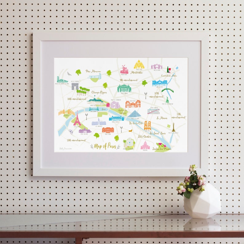 May include: A colorful illustrated map of Paris, France, with landmarks and neighborhoods labeled. The map is framed in a white frame and is hanging on a white pegboard wall.