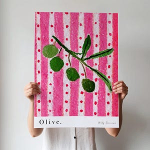 Puede incluir: Una pintura de acuarela de una rama de olivo con aceitunas verdes sobre un fondo a rayas rosa y blanco con lunares blancos. La pintura est&aacute; titulada "Olive." y firmada "Holly Franzen".
