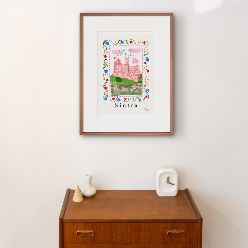 May include: A framed illustration of a pink castle with a blue sky and white clouds. The castle is surrounded by a colorful floral border. The text "Sintra" is written below the castle.