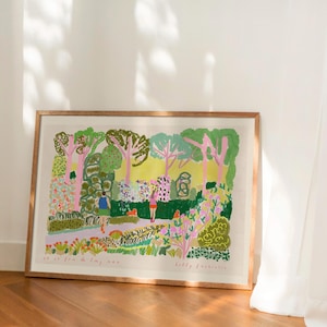 May include: A framed art print depicting a colorful park scene with trees, flowers, and figures. The artwork features a variety of green, pink, and yellow hues, with visible brushstrokes. The print is displayed in a wooden frame and rests on a wooden floor.