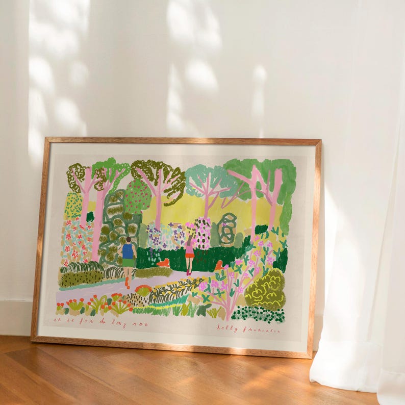 May include: A framed art print depicting a colorful park scene with trees, flowers, and figures. The artwork features a variety of green, pink, and yellow hues, with visible brushstrokes. The print is displayed in a wooden frame and rests on a wooden floor.