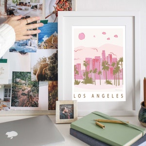 K&ouml;nnte beinhalten: Ein wei&szlig;er gerahmter Druck mit einer rosa und gr&uuml;nen Illustration der Skyline von Los Angeles. Der Druck hat den Text "LOS ANGELES" in goldenen Buchstaben unten.