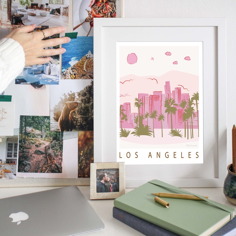 K&ouml;nnte beinhalten: Ein wei&szlig;er gerahmter Druck mit einer rosa und gr&uuml;nen Illustration der Skyline von Los Angeles. Der Druck hat den Text "LOS ANGELES" in goldenen Buchstaben unten.