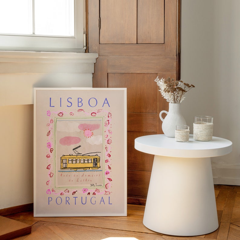 May include: A vintage-style poster with a yellow tram in front of a pink and blue sky. The poster says "LISBOA" in blue letters and "PORTUGAL" in blue letters at the bottom. The poster is framed in white and is sitting on a wooden floor.