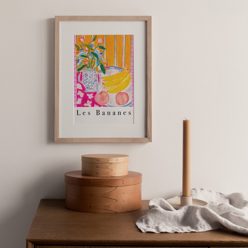 Op de afbeelding: Een ingelijste print van een stilleven met een vaas met bloemen, bananen en perziken. Het schilderij is getiteld "Les Bananes" en hangt aan een witte muur boven een houten tafel met twee ronde houten dozen en een kaarsenstandaard.